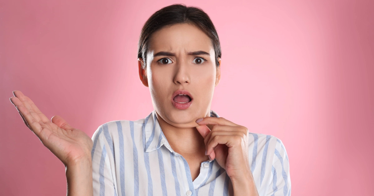 A woman with a surprised or concerned expression touches her chin, gesturing thoughtfully, against a pink background, hinting at Kybella treatment in Oak Brook, IL.